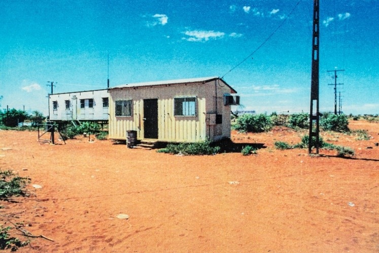 A grainy photo of a yellow and silver demountable, surrounded by red dirt, green bush, and blue sky.