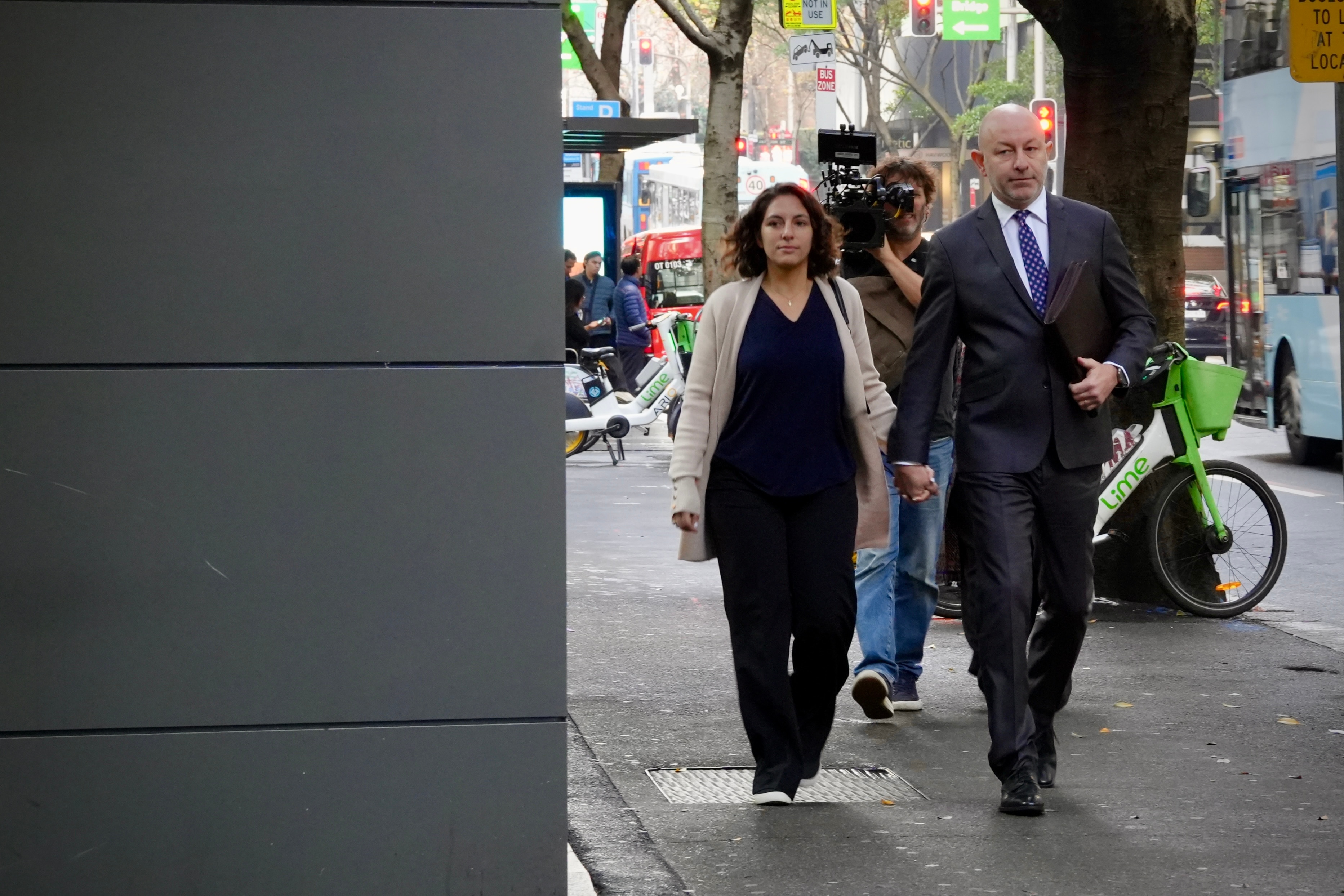 Mr Ostrovsky in a black suit and blue tie walking next to a woman in a black dress with a news camerman behind them.