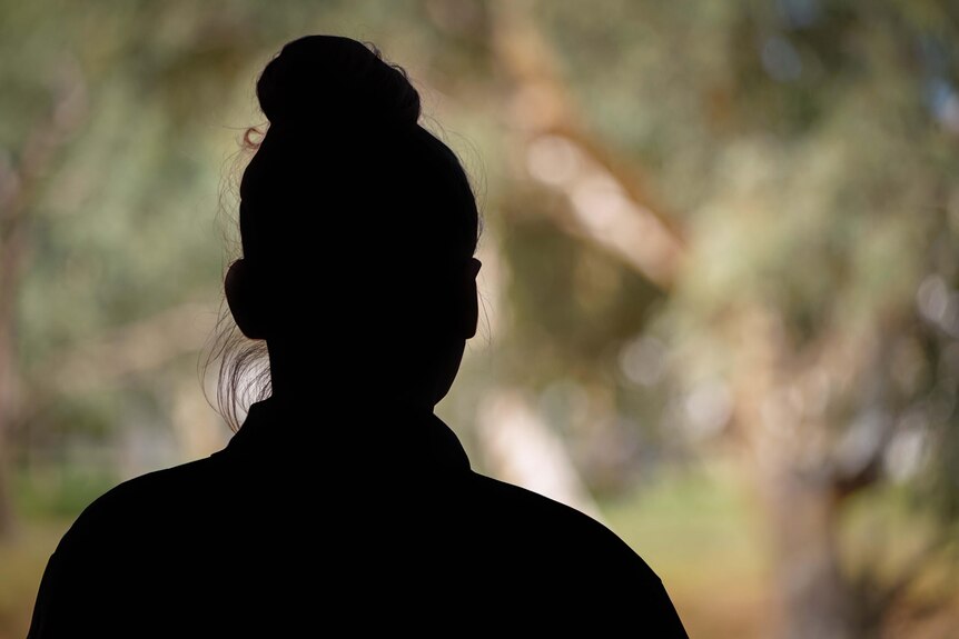 A silhouette of a woman's head. Her hair is in a bun and she is outside, looking out onto bush.