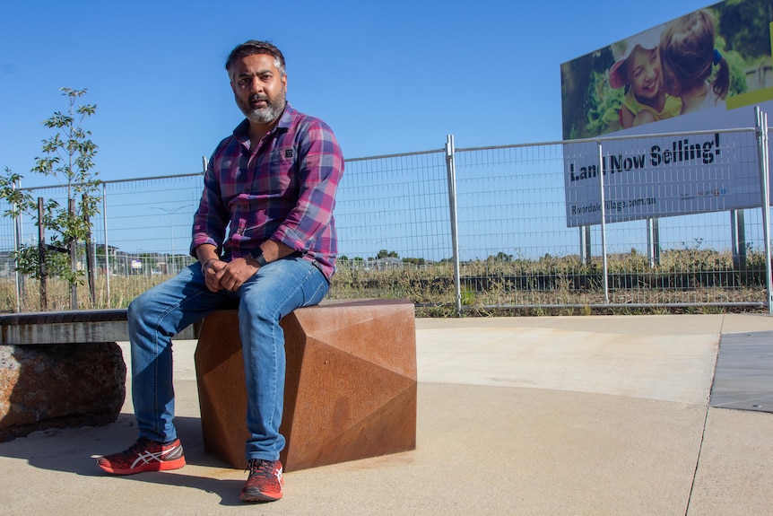 A man sits on a bench seat in front of a sign that says "land now selling"