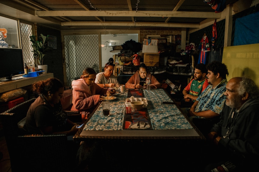 Seven people sit around a table on a covered balcony at night. Some are eating. Boxes are stacked against a back wall.