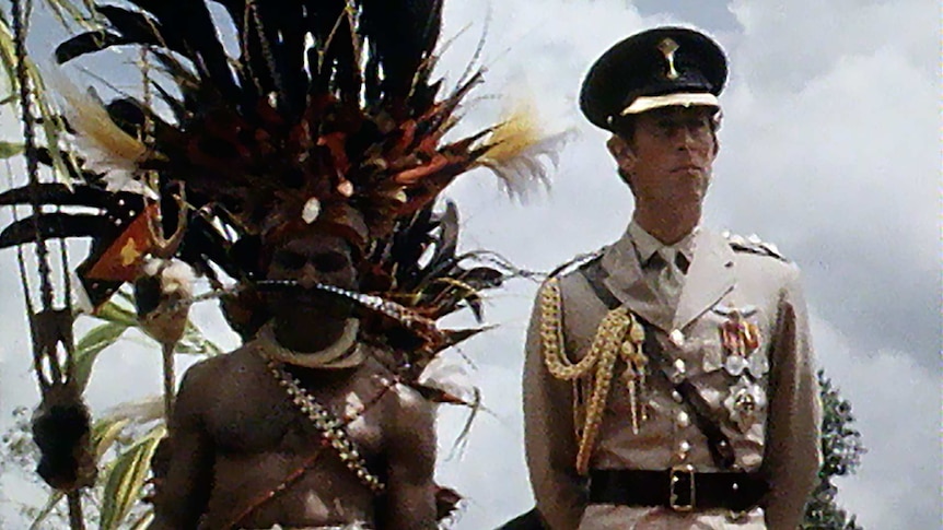Prince Charles in military uniform speaks to a crowd standing next to indigenous PNG man in feathered headdress.