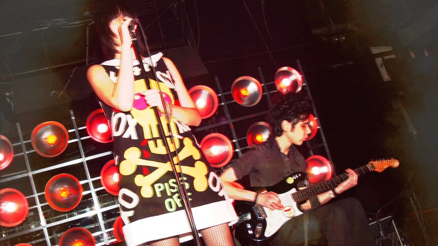 Karen O wears a designer dress and performs live with guitarist Nick Zinner in front of a bank of flash bulb lighting