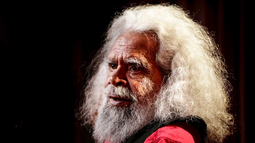 Jack Charles sits looking across to the side, with lighting illuminating his white hair.