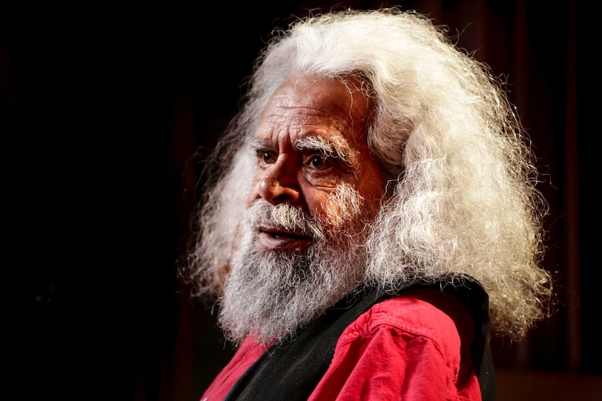 Jack Charles sits looking across to the side, with lighting illuminating his white hair.