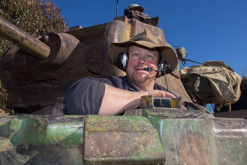 A man wearing a floppy felt hat over earphones and a headset in the cabin of a tank