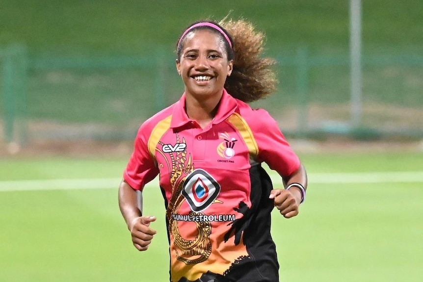A smiling woman with thick, curly, long dark hair tied back runs towards the camera, wearing a pink cricket shirt.