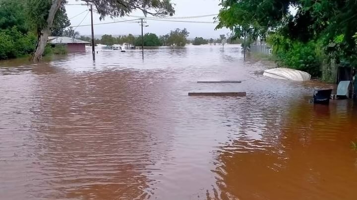 A heavily flooded road between houses in a remote community, with a small boat and a few planks floating in the water.
