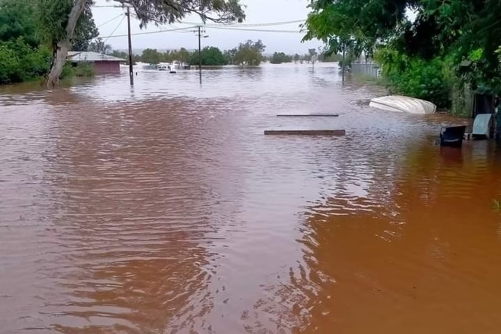 A heavily flooded road between houses in a remote community, with a small boat and a few planks floating in the water.