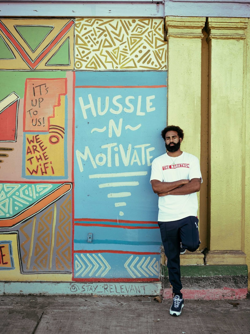A man stands in front of a painted wall that says hussle n motivate.