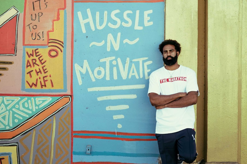 A man stands in front of a painted wall that says hussle n motivate.