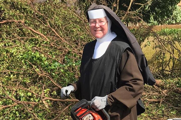 Sister Margaret Ann, dressed head to toe in her nun's habit, smiles while holding a chainsaw in front of fallen trees.