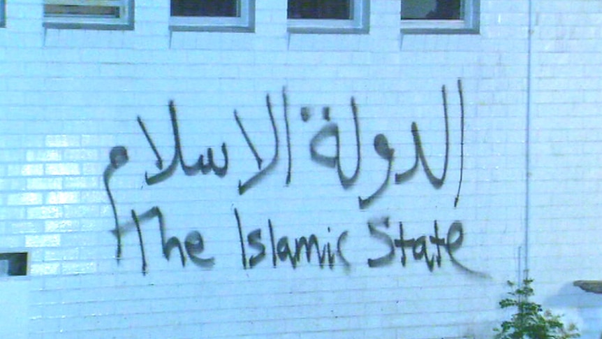 The words 'the Islamic State' scrawled in big spray-painted Arabic and English letters on a white brick wall.