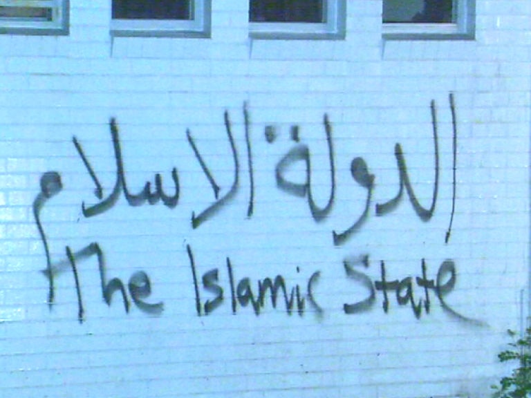 The words 'the Islamic State' scrawled in big spray-painted Arabic and English letters on a white brick wall.