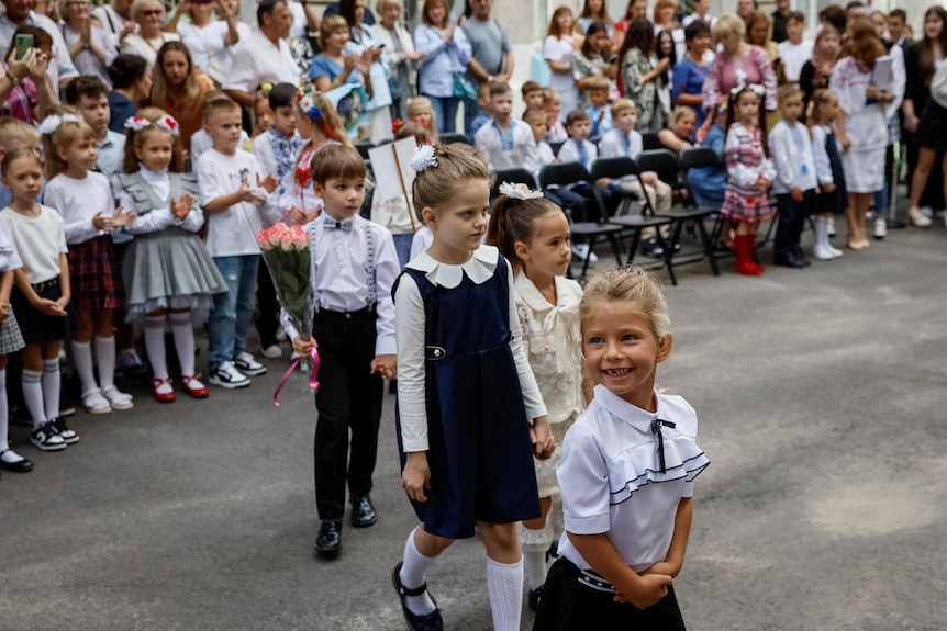 A large group of school-age children wearing nice clothing such as dresses have gathered for a ceremony