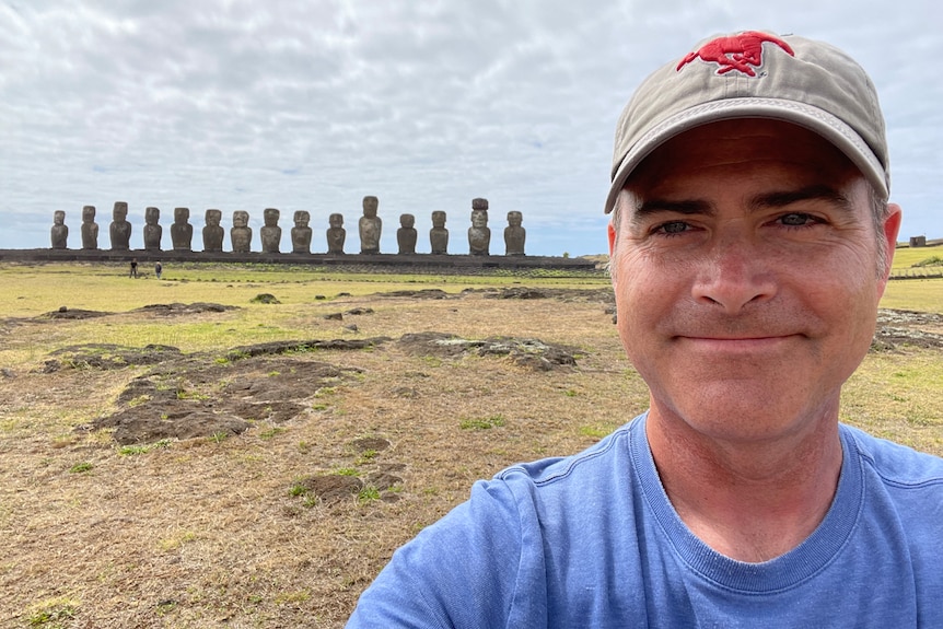 A man wearing a hat and blue shirt takes a selfie with huge monoliths in the distant background