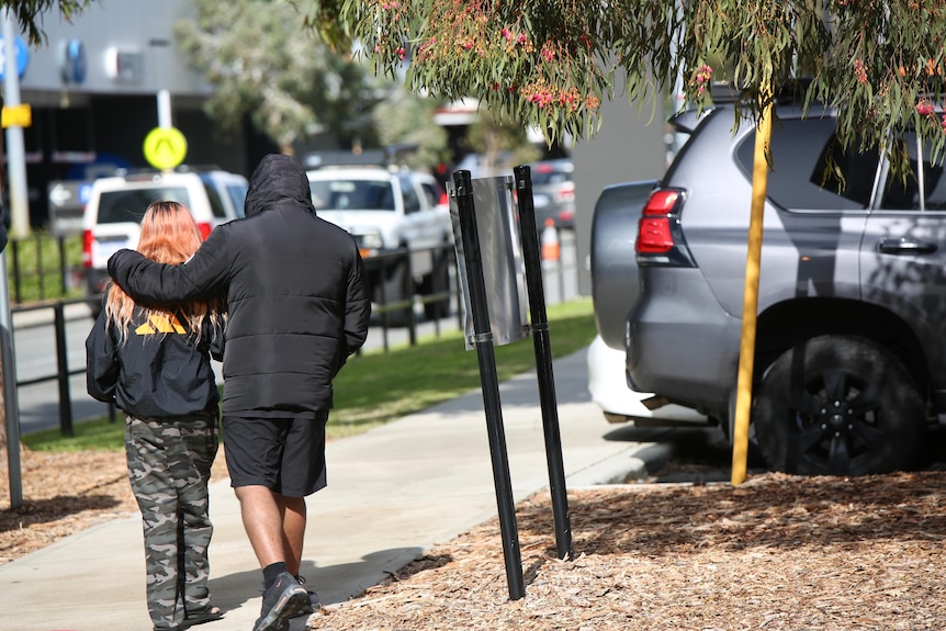 A woman with pink hair and cargo shorts walks away while a man in a black puffer jacket has his arm around her.