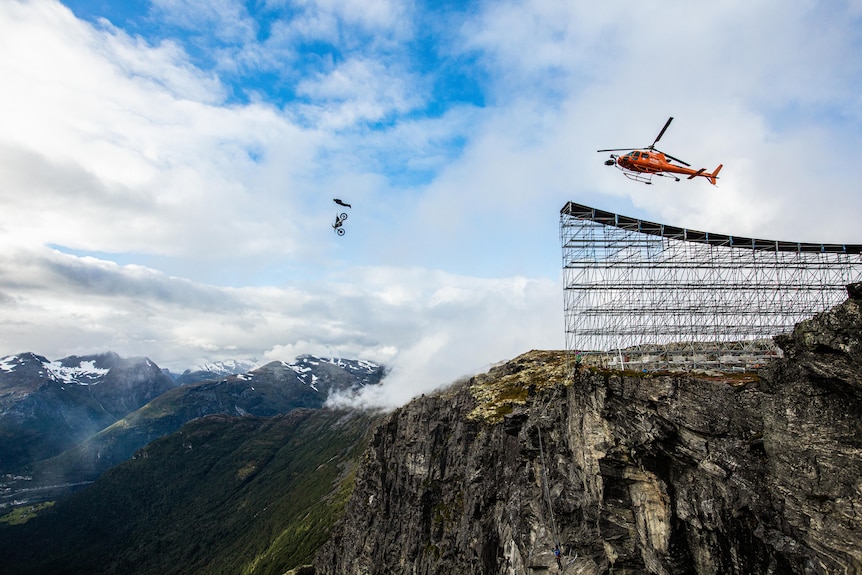 A helicopter flies above the mountains. There is a ramp made of scafold and a man is suspended above a motorbike high above.