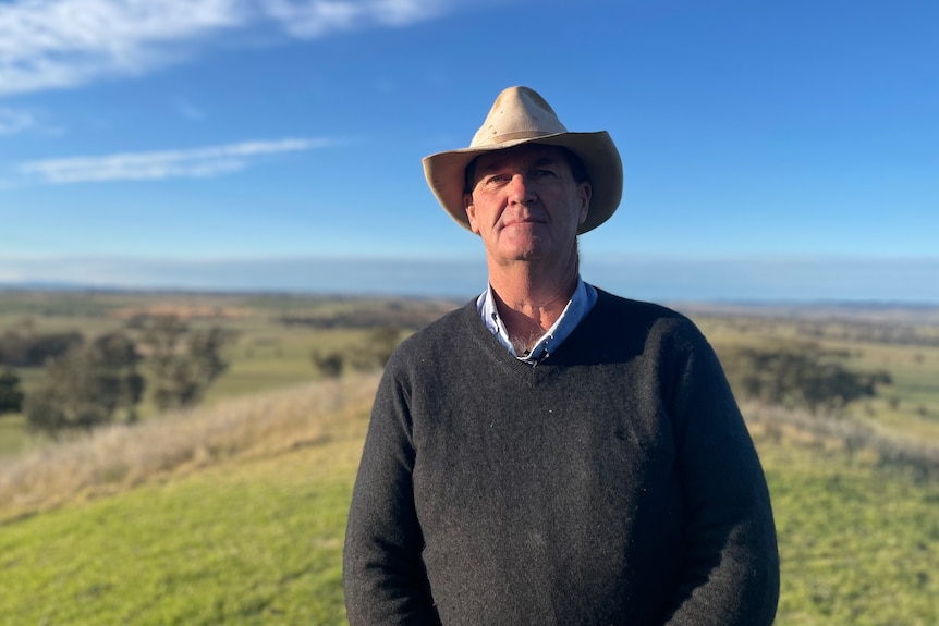 A farmer wearing a hat and v-neck jumper standing in a paddock