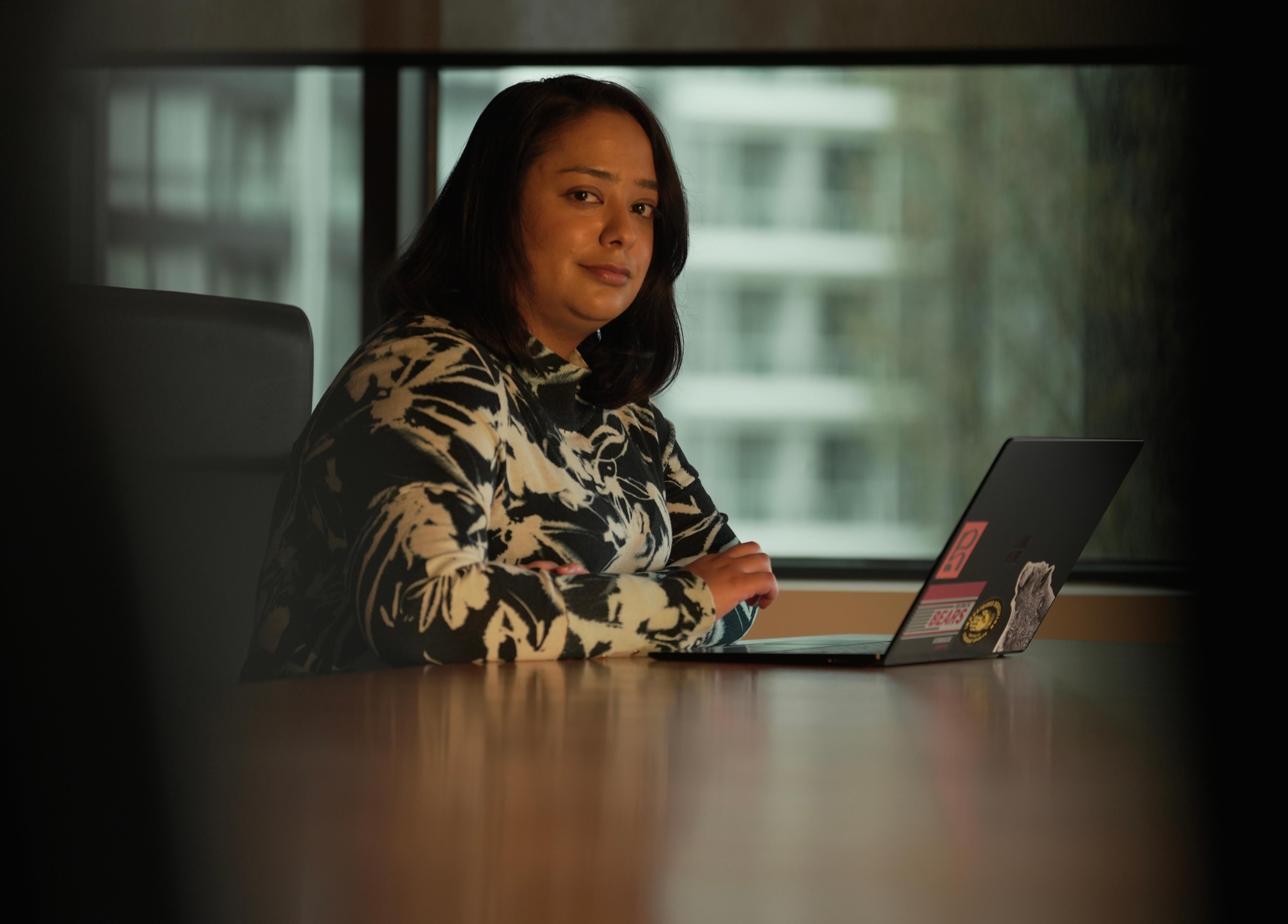 Trisha Jha stares at the camera, in a dimly lit office, with a laptop open in front of her.