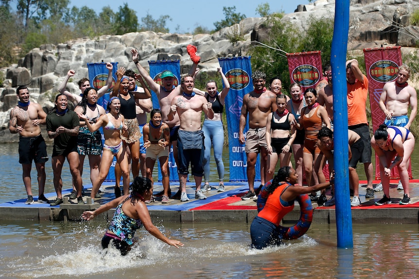 A group of Survivor contestants standing in front of coloured flags cheer at two women in a creek