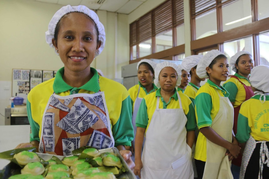 A woman with a tray of green baked foods in the foreground with a group of women behind her.