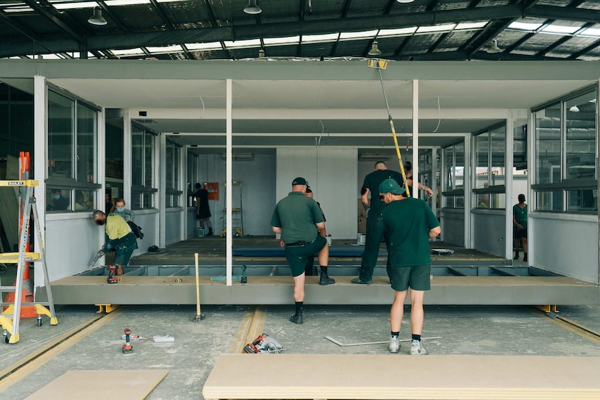 Prison inmates wearing green pain and fabricate a classroom inside a warehouse.