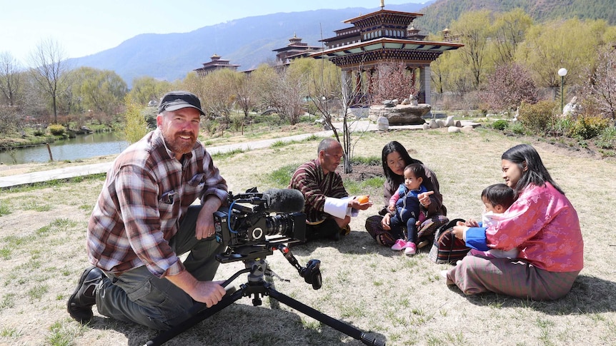 Smith with camera filming twins and their family sitting on grass outside with temples in background.