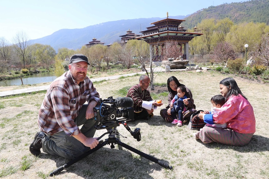 Smith with camera filming twins and their family sitting on grass outside with temples in background.
