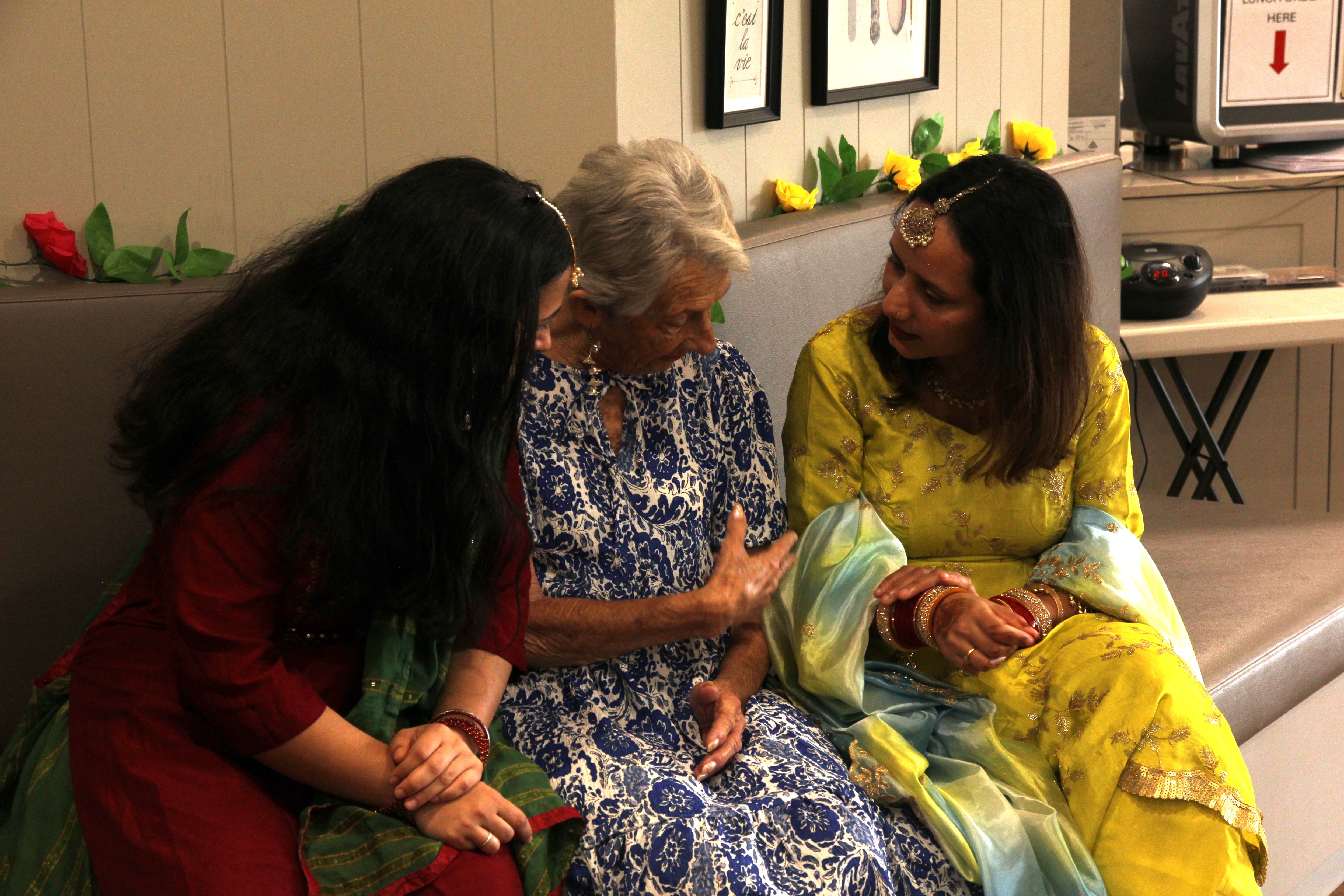 Dos mujeres indias con saris tradicionales se sientan a cada lado de una mujer blanca mayor con un vestido mientras acaricia la tela.