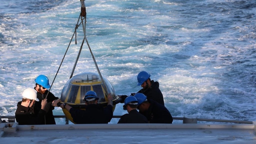 Men in hard hats prepare to lower large buoy into ocean.