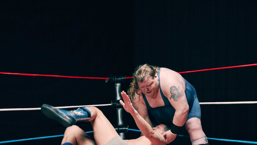 Large wrestler flipping over his smaller competitor, held by the neck.