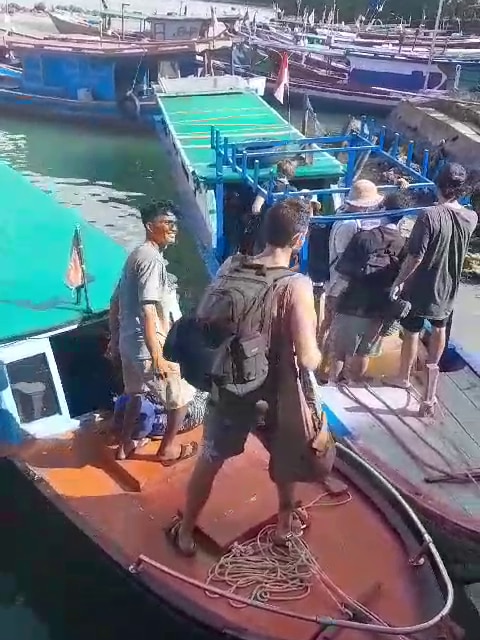 A group of people file onto two fishing boats, one man smiles while helping them on