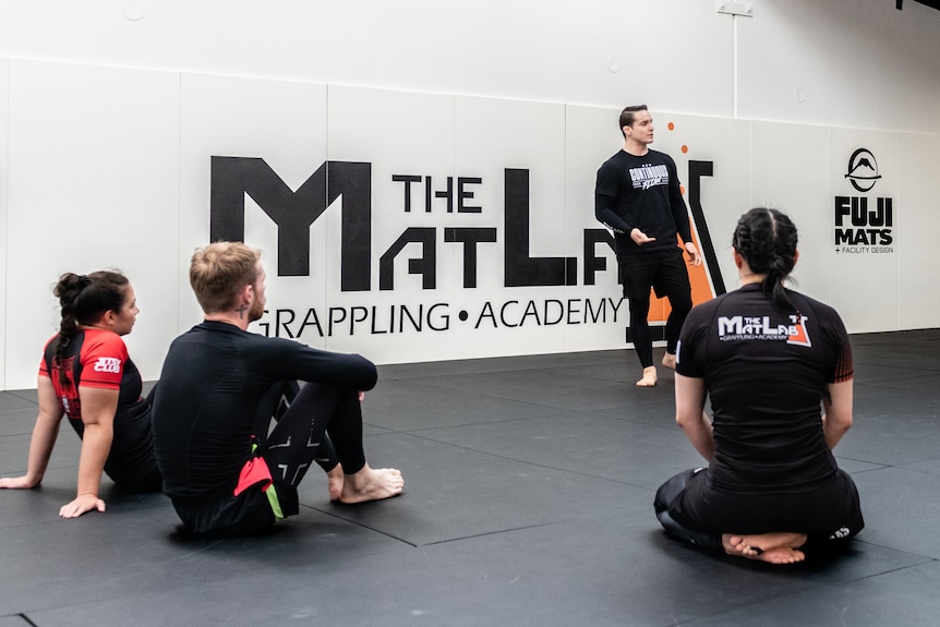 People sit on a mat in a gym and listen to an instructor.