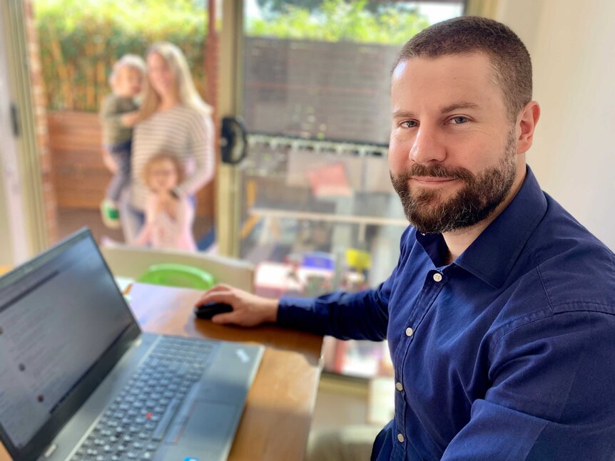 Adam Hart sits at a laptop computer at home with wife and two children blurred in the background.
