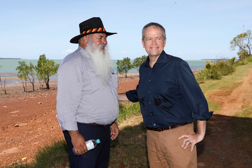 The two men smile as they stand on a dirt road alongside water