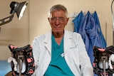Older male doctor sitting in a clinic room, wearing his scrubs and lab coat. 