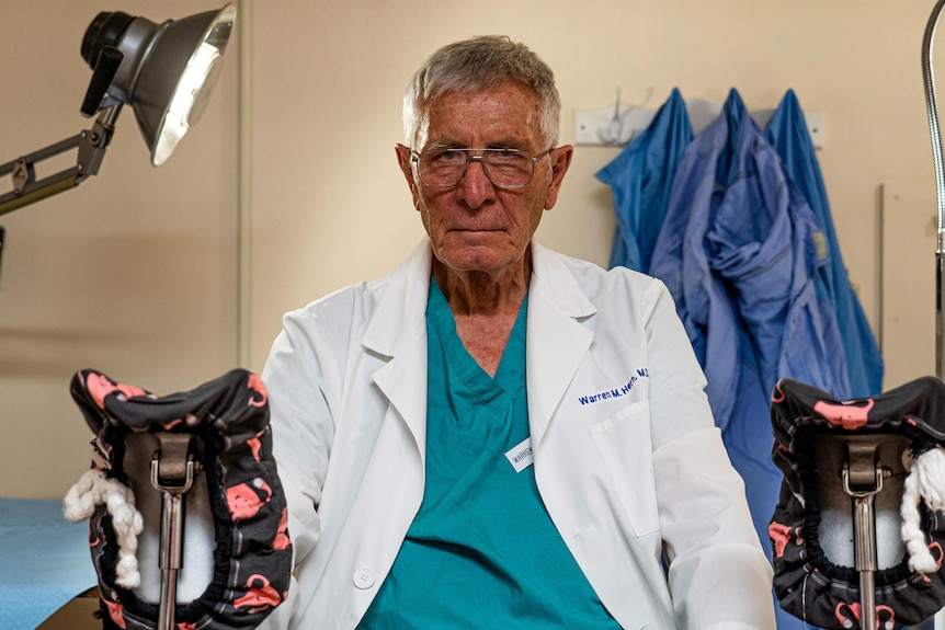 Older male doctor sitting in a clinic room, wearing his scrubs and lab coat. 