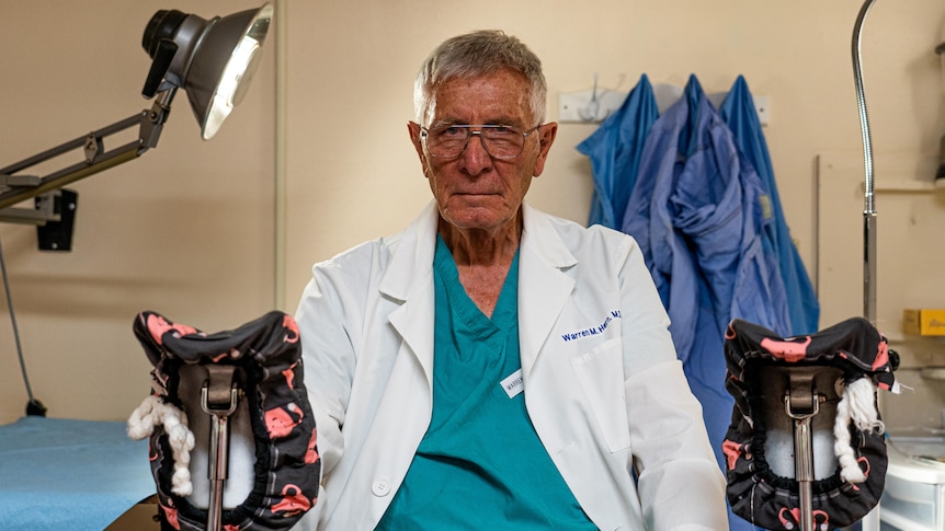 一位年长的医生坐在诊室里，穿着磨砂膏和实验室外套。 