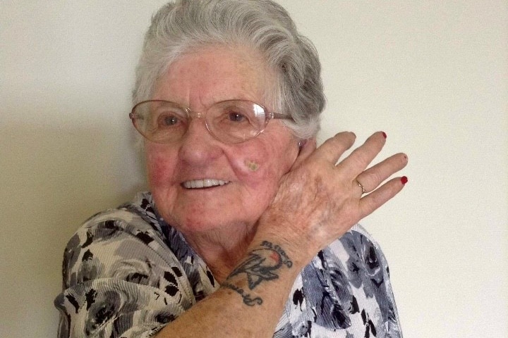 An elderly woman smiles for the camera and shows off her Sydney Swans premiership tattoo on her right forearm