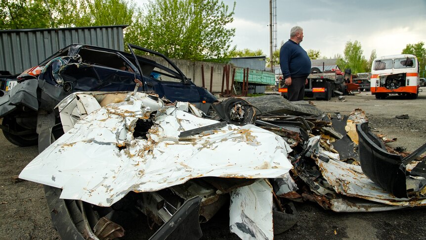 The smashed-up wreckage of two small sedans sits in a carpark with other buses and trucks. A man stands nearby