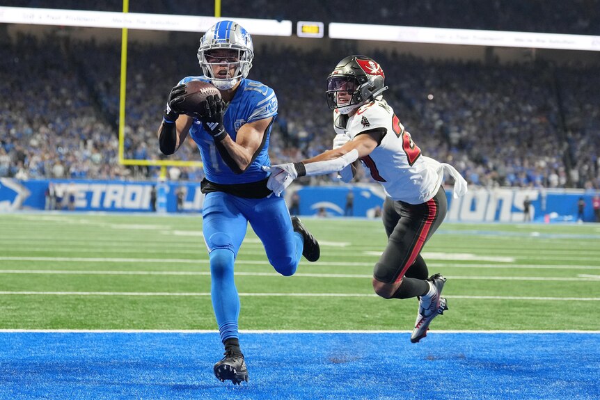 A Detroit Lions American footballer holds the ball as he steps in the end zone for a touchdown next to a defender.