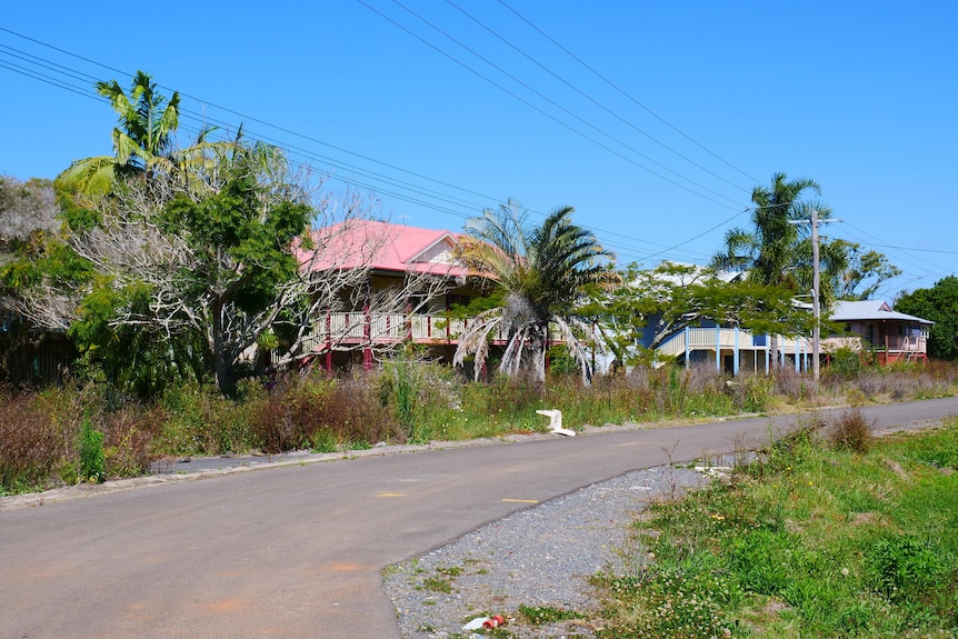 two storey Queenslander style homes with overgrown weeds along verge, plastic chair on road