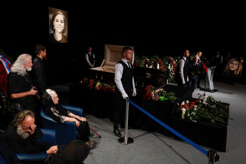 Mourners surround a casket decorated with flowers. 