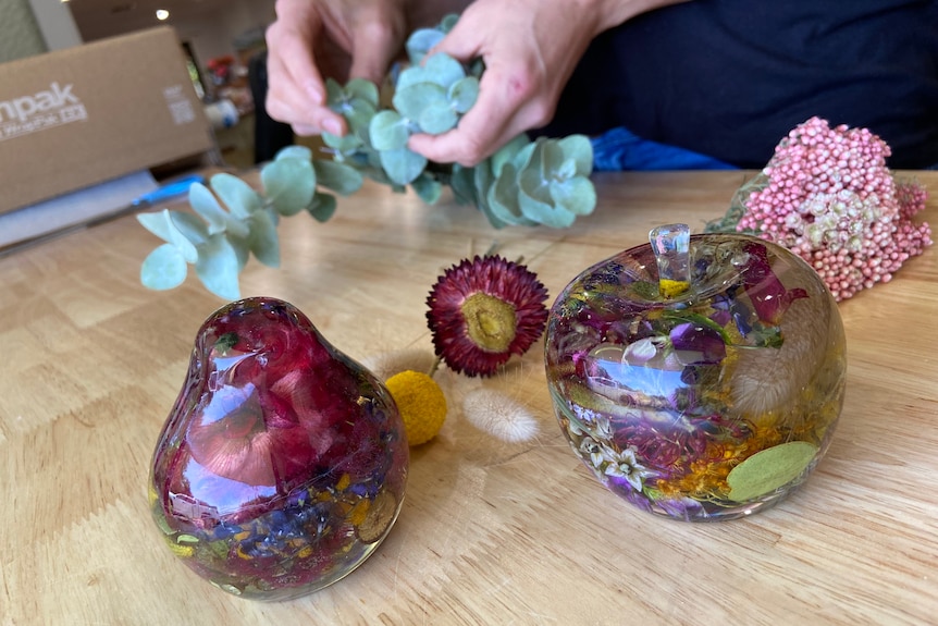 An apple and bird resin ornaments filled with flowers and a woman's hands holding foliage in background.