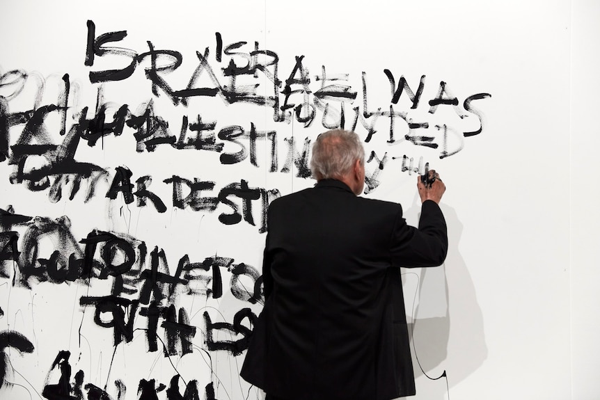 Artist Mike Parr painting text onto a white gallery wall with black paint, while his eyes are closed.