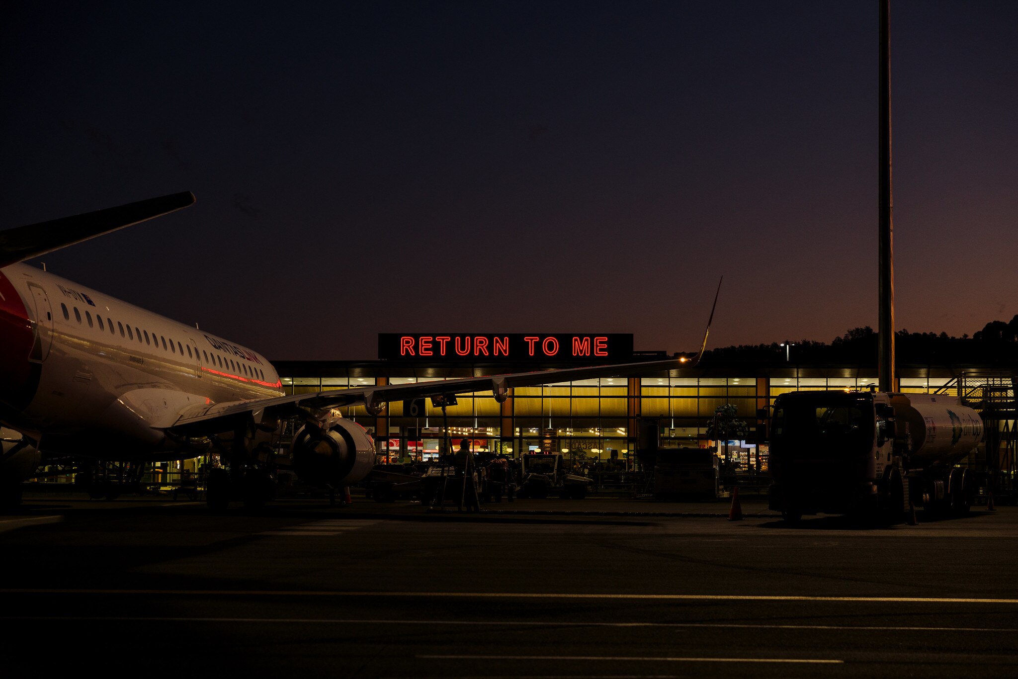 A red neon sign says 'return to me' at Hobart airport, a aeroplane is also in the picture