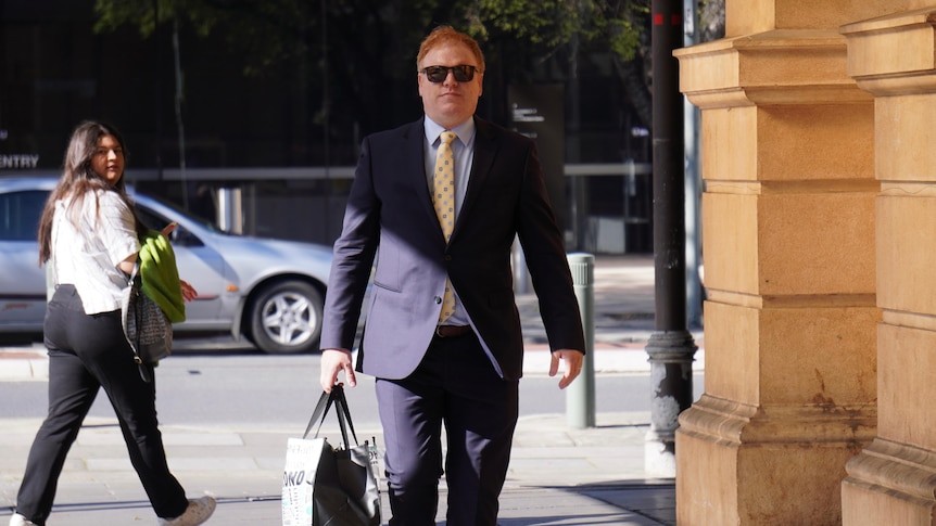 A man in a suit and glasses carrying a bag walks along a sidewalk next to a sandstone-colored building