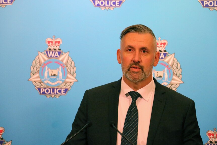 A police officer in a suit named Troy Casey speaks in front of a logo that says 'WA Police'.