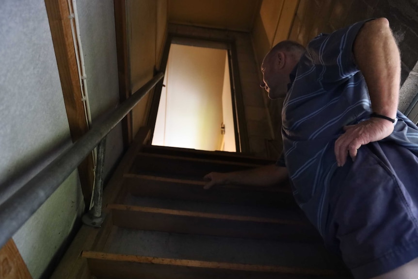 A man dressed in a blue shirt and shorts on a step of a ladder leading to a light filled door